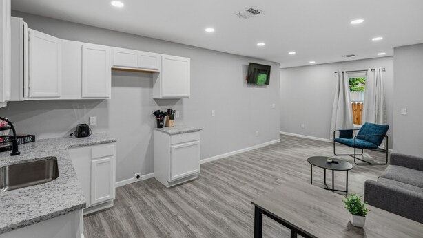 A modern kitchen with white cabinets, a granite countertop, and a coffee machine. There is a dining area with a gray sofa and a small table. - Spacious 3BR Retreat Near Downtown River Walk (San Antonio)