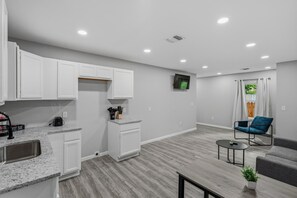 A modern kitchen with white cabinets, a granite countertop, and a coffee machine. There is a dining area with a gray sofa and a small table. - Spacious 3BR Retreat Near Downtown River Walk (San Antonio)