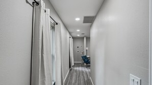 A narrow hallway with white walls and a wooden floor, leading to a room with a blue chair and a small table. - Spacious 3BR Retreat Near Downtown River Walk (San Antonio)