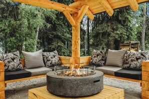 A wooden outdoor seating area with a fire pit, surrounded by a forest.