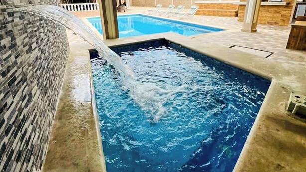 A swimming pool with a water feature, surrounded by a tiled wall and a view of a building with a blue roof. - Villas cloud 1 (Santo Domingo)