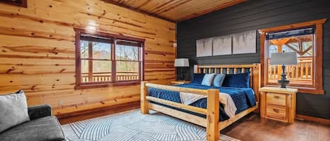A log cabin bedroom with a bed, a nightstand, a lamp, a ceiling fan, and a window with a view.
