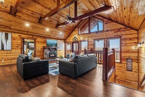A log cabin interior with a fireplace, two sofas, a ceiling fan, and large windows. - 8-Bedroom Cabin with Indoor Pool, Mini Golf & Hot Tub, Sleeps 30 (Sevierville)