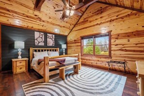 A wooden cabin bedroom with a bed, bedside tables, a ceiling fan, and a window with a view of greenery.