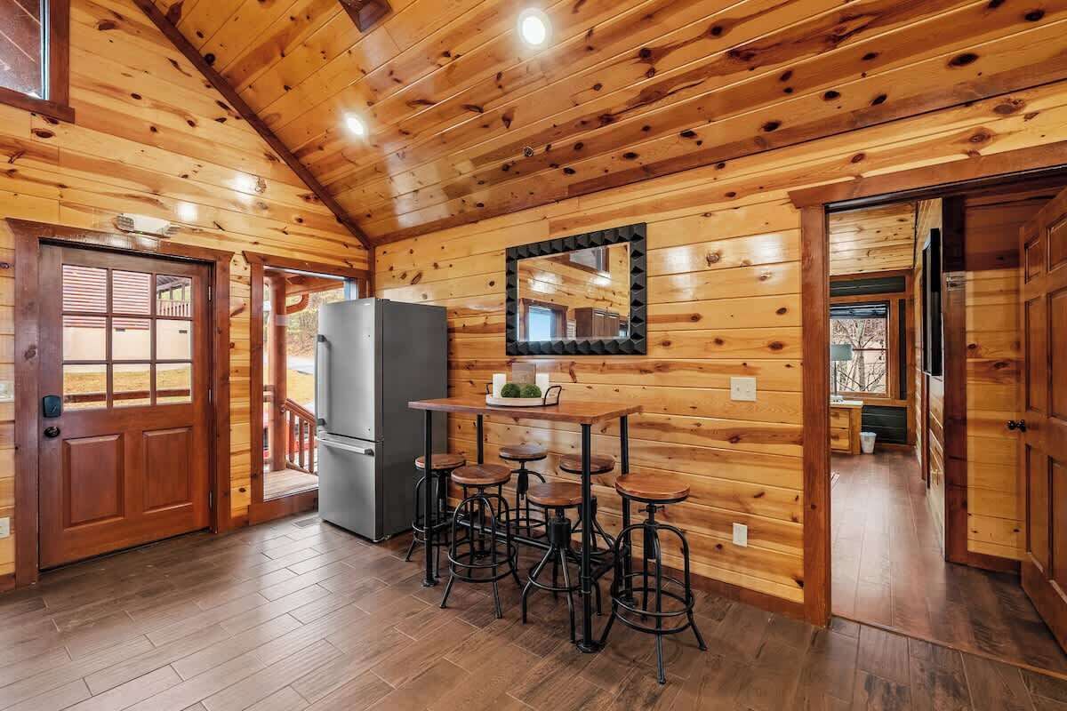 A wooden interior with a kitchen area featuring a refrigerator, a table with stools, and a mirror on the wall.
