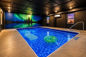 An indoor swimming pool with a view of the northern lights on the wall, a floating lounge, and a window with a sunset view. - 8-Bedroom Cabin with Indoor Pool, Mini Golf & Hot Tub, Sleeps 30 (Sevierville)