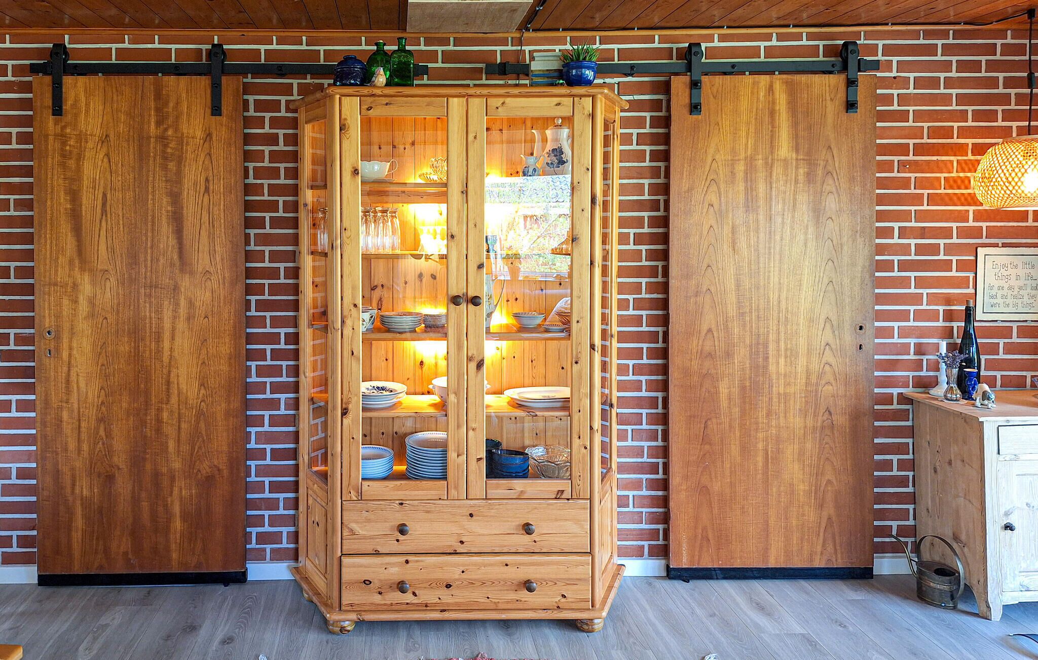 A wooden cabinet with glass doors against a brick wall, containing dishes and glassware.