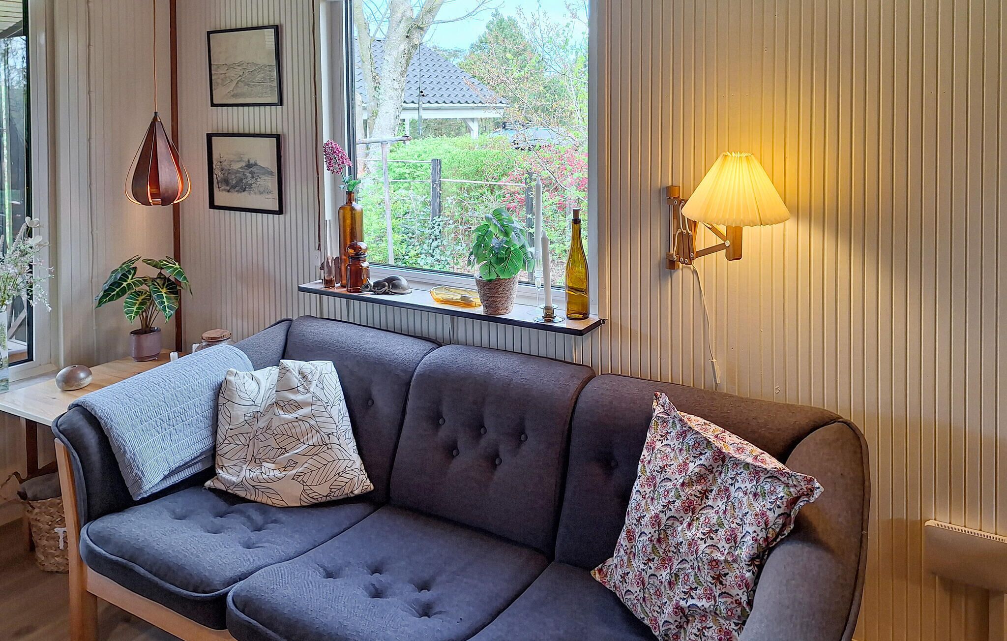 Un salon chaleureux avec un canapé gris, deux coussins à motifs, une petite table agrémentée d’objets décoratifs, une fenêtre donnant sur un paysage verdoyant et une lampe fixée au mur.
