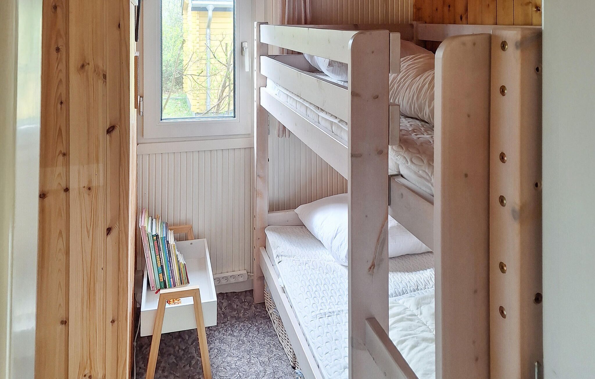 Ein Schlafraum mit Etagenbetten, einem kleinen Tisch und Büchern, einem Fenster mit Blick auf Bäume und Holzwänden.