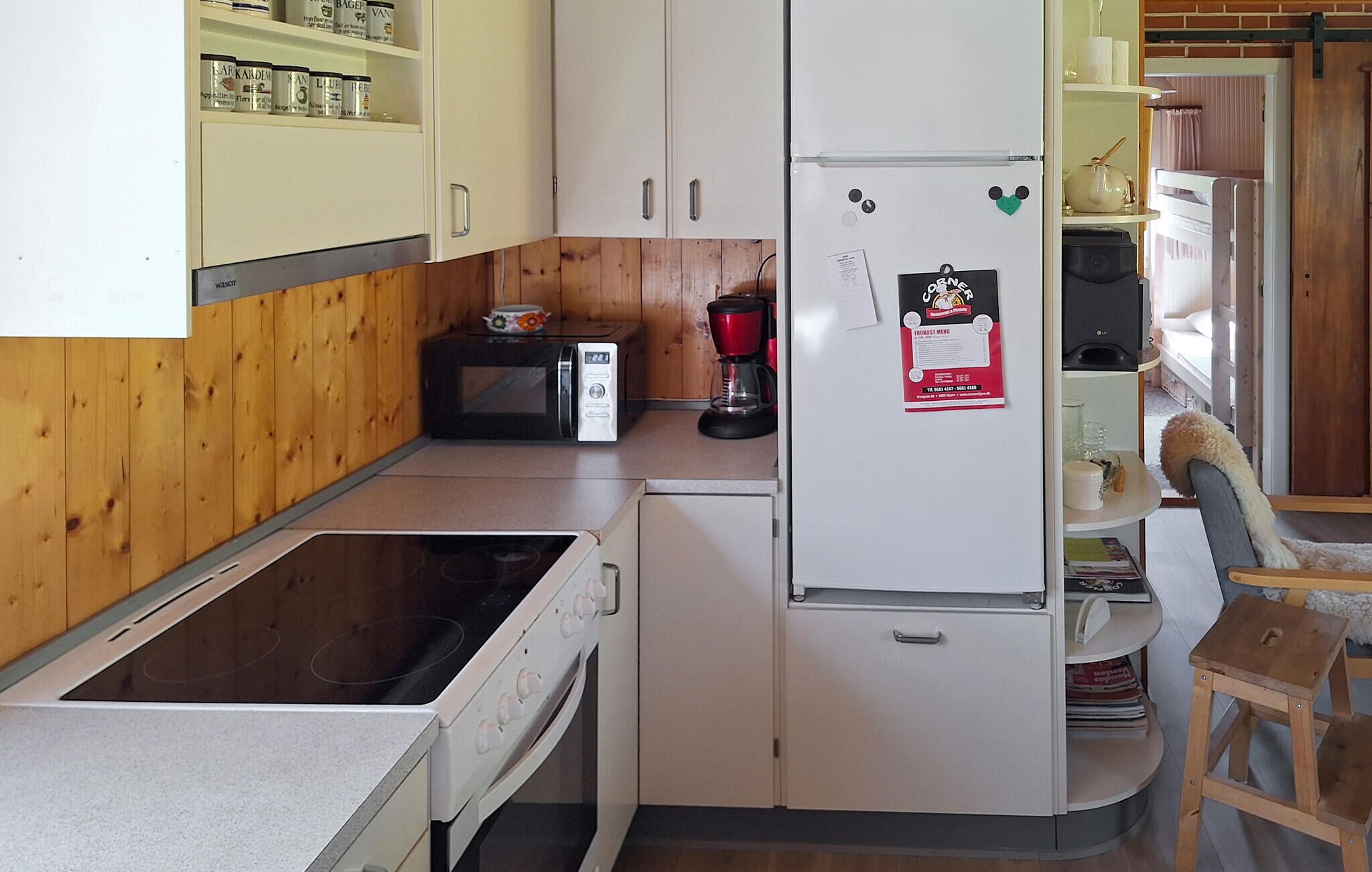 A compact kitchen with a stove, microwave, and refrigerator. There is a wooden cabinet with jars and a coffee maker on the counter.