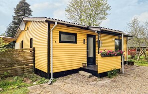 A yellow cabin with a black door, number 6, a small window, and a flower box with pink flowers.