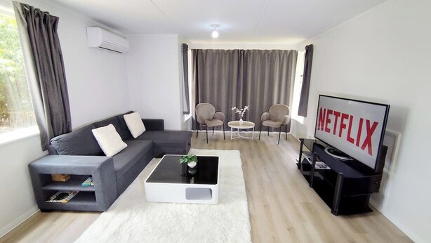 A modern living room with a grey sofa, a white coffee table, a TV, and a dining area with chairs. - 3-Bedroom House +Private yard+sliding gate (Hamilton)