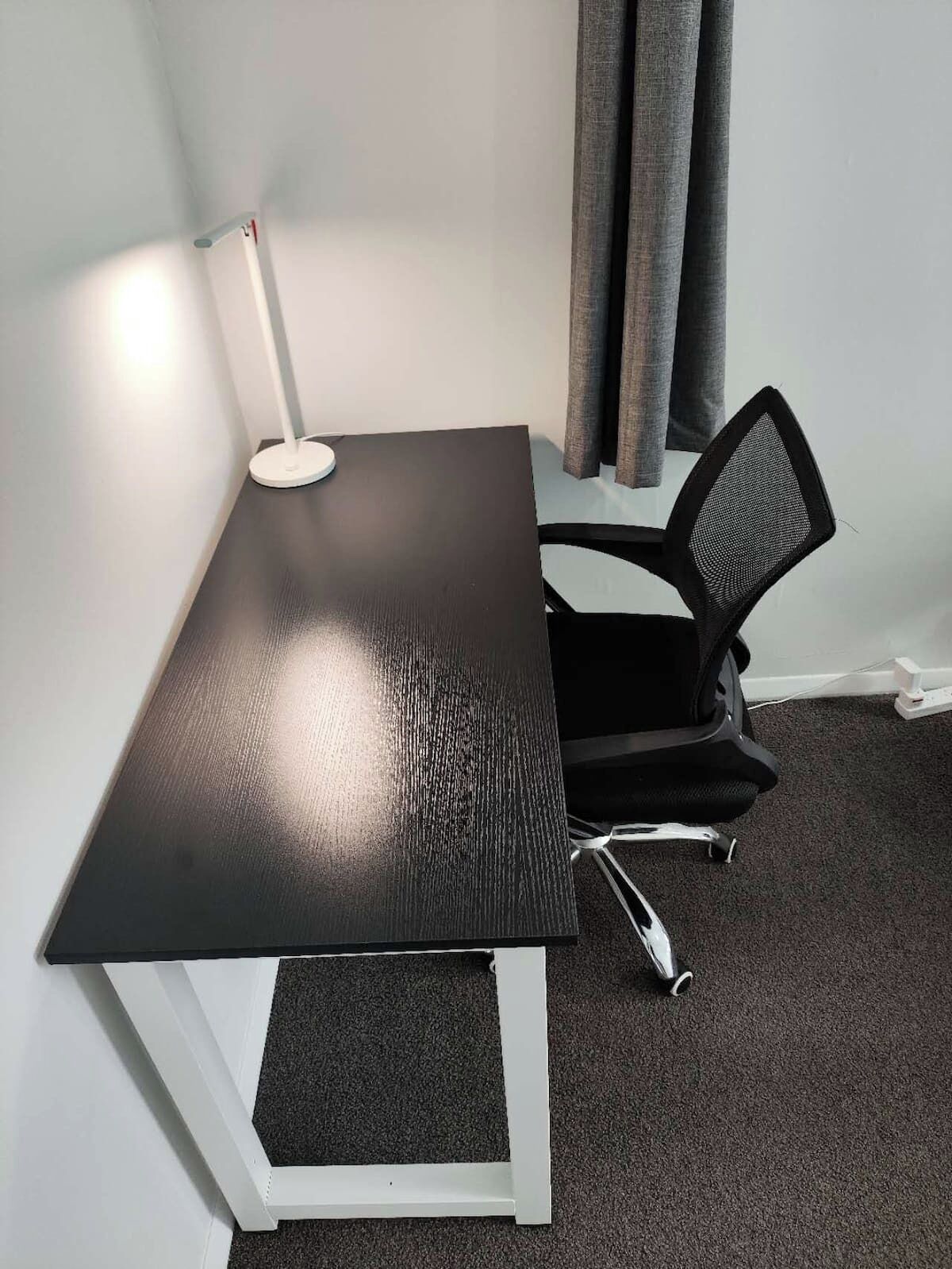 Une petite table de couleur sombre, dotée d’un cadre blanc, une chaise de bureau noire et un lampadaire blanc.