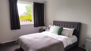 A neatly made bed with a grey headboard, white linens, and a green pillow. A small bedside table with a lamp and a clock is visible. The room has a window with black curtains, and the walls are painted white. - 3-Bedroom House +Private yard+sliding gate (Hamilton)