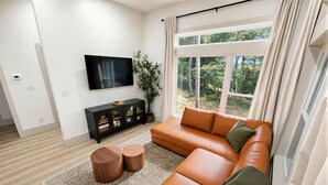 A modern living room with a sectional sofa, a flat-screen TV, a black entertainment unit, and a large window with a view of trees. - Waterfront Luxury - Hot Tub, Game Room, Kayaks, Best View (Cadiz)