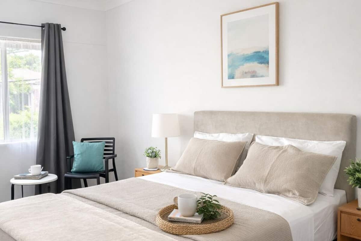 Une chambre à coucher bien rangée, avec un grand lit, une table de chevet, une chaise et une fenêtre avec des rideaux.