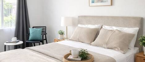 A neatly arranged bedroom with a large bed, a bedside table, a chair, and a window with curtains.