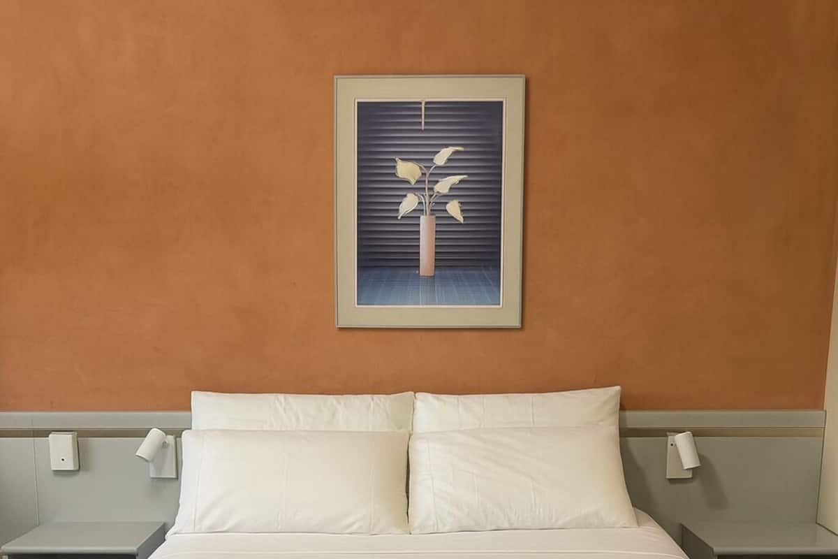 Un lit bien fait, avec des draps blancs, une couverture bleue et deux serviettes enroulées. Un cadre avec une photo d’une plante est accroché au mur, au-dessus du lit.