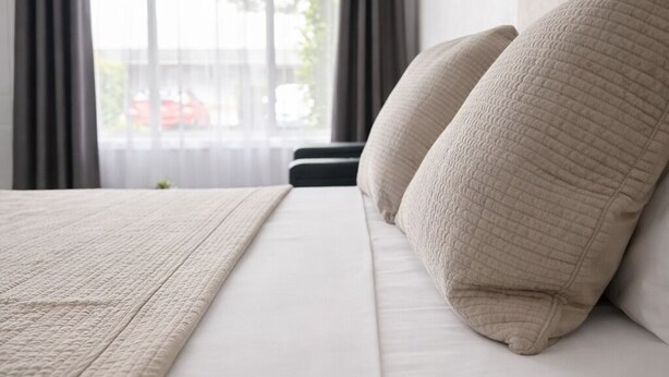 A neatly made bed with a textured blanket and pillows, a window with curtains, and a view of the outside. - Banksia Motel Bass Hill (Bass Hill)
