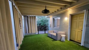 Uma área externa coberta com grama sintética verde, um sofá branco e uma mesa redonda pequena.