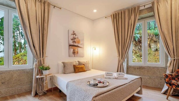 A modern bedroom with a large bed, a bedside table with a lamp, and a view of greenery through the windows. - Rustic & Modern Stone House by Host Wise (Pedroso)