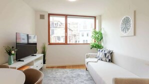 A modern living room with a flat-screen TV, a sofa, a dining table, and a large window with a city view.