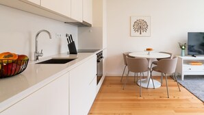 A modern kitchen with white cabinetry, a stainless steel sink, and a fruit bowl. There is a dining area with a round table and four chairs. A flat-screen TV is mounted on the wall.
