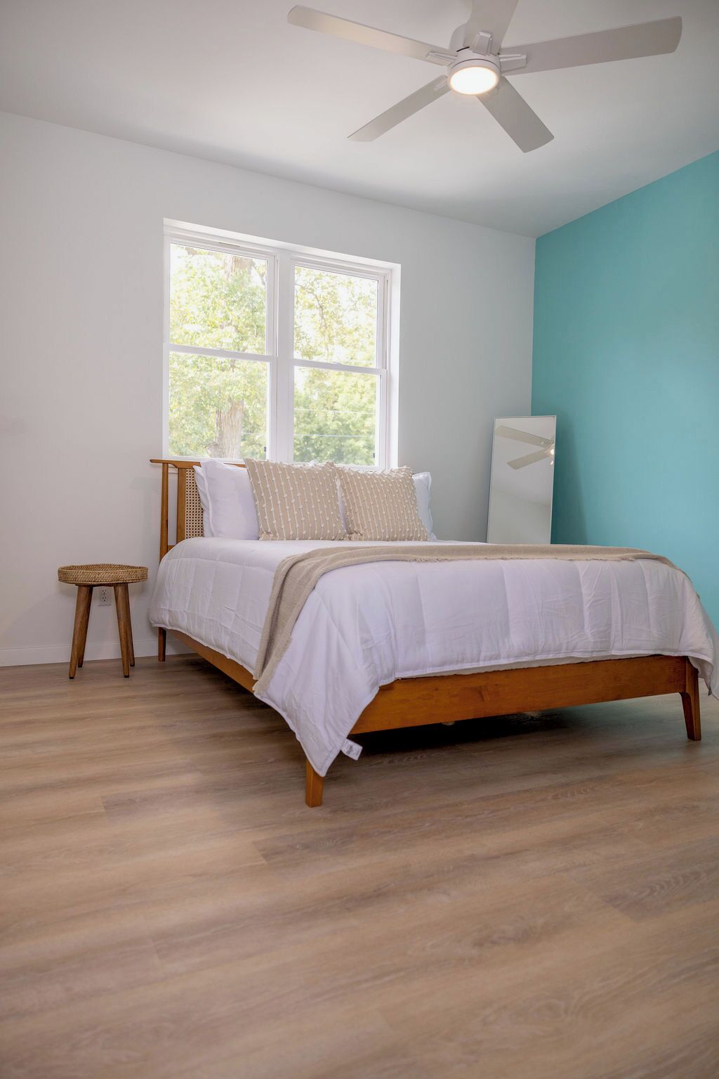 Un dormitorio con una cama de madera, un taburete pequeño, un ventilador de techo y una ventana con vistas al verdor.