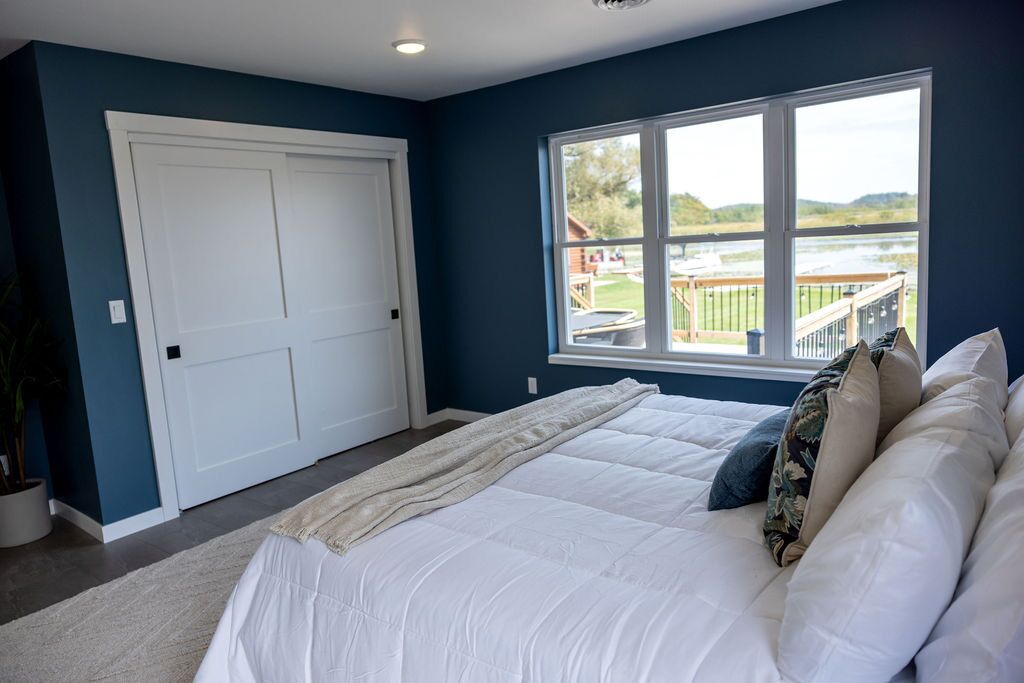 A bedroom with a large bed, a window with a view of a deck and water, and a white door.