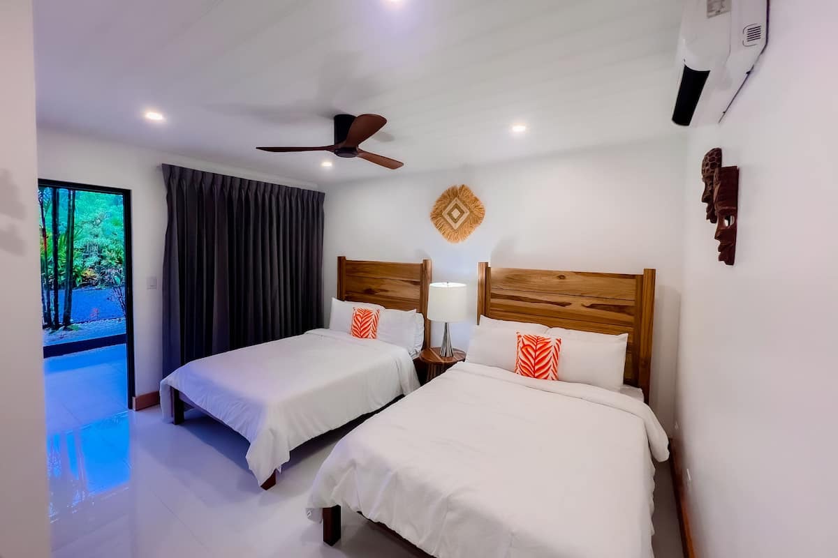 A hotel room with two single beds, a ceiling fan, an air conditioning unit, a nightstand with a lamp, and a view of greenery through a window.