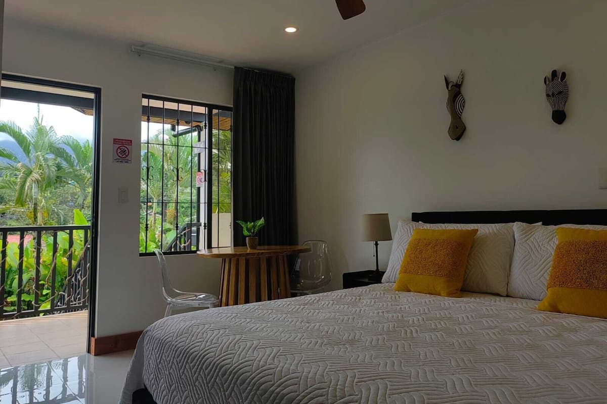A hotel room with a bed, a desk, a chair, a lamp, and a view of the outdoors.