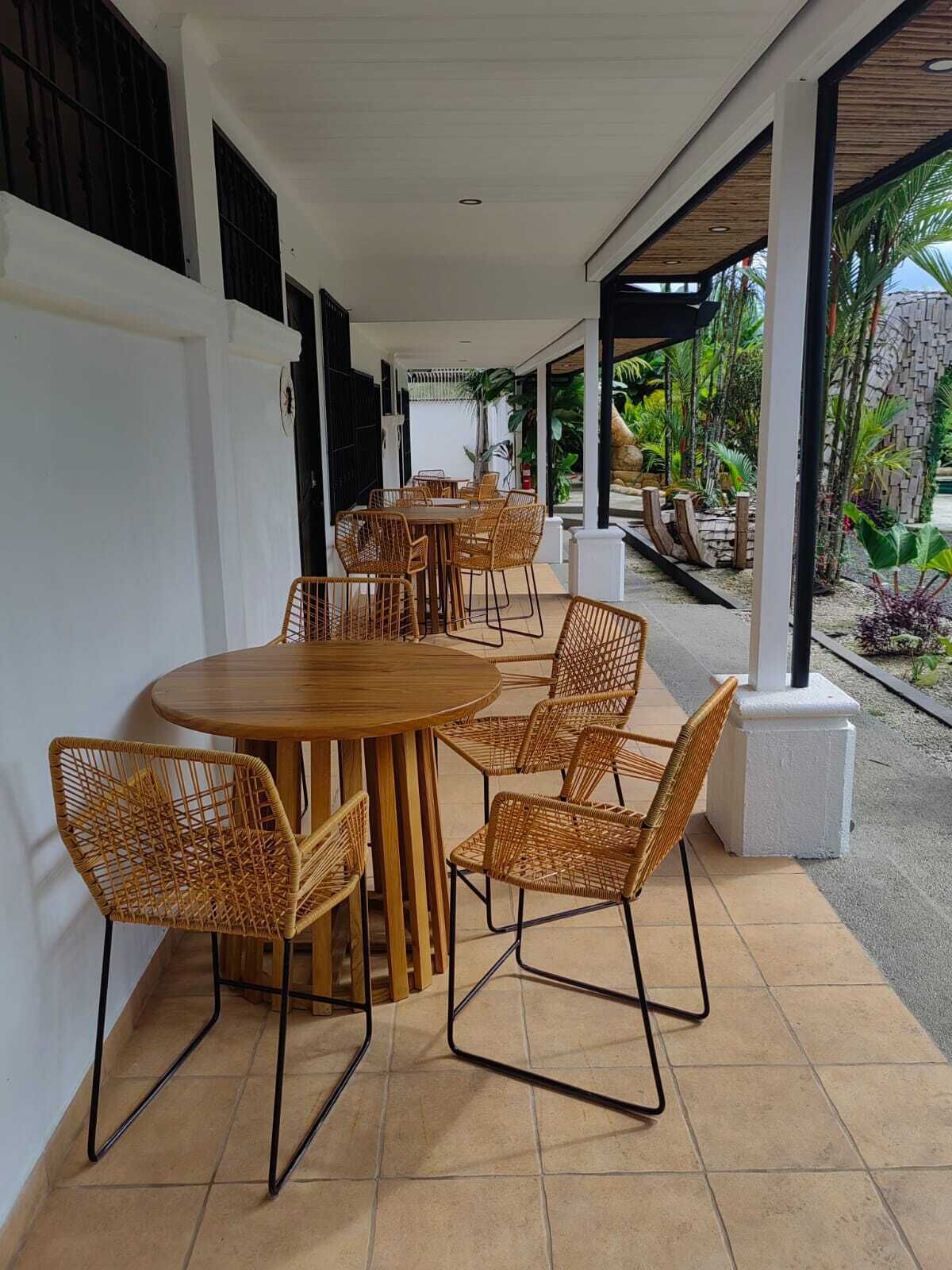An outdoor patio area with a round wooden table and wicker chairs, surrounded by white walls and a tiled floor.