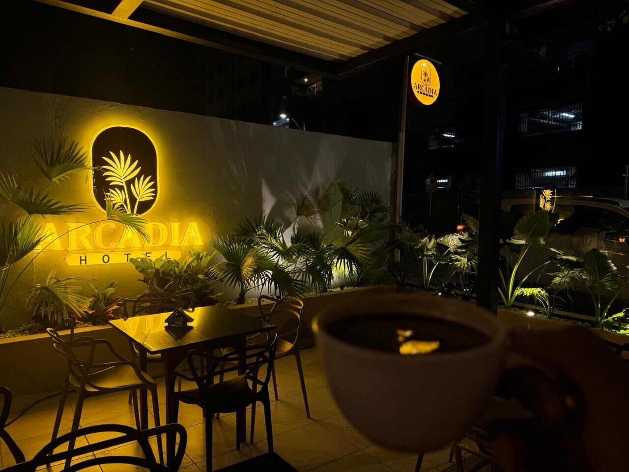 A nighttime view of the Arcadia Hotel's outdoor seating area with a table and chairs, surrounded by potted plants and a sign with the hotel's name.