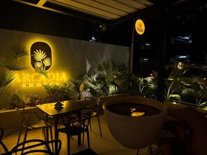 Een nachtzicht van het buitenzitgedeelte van Hotel Arcadia, met een tafel en stoelen, omgeven door potplanten en een bord met de naam van het hotel.