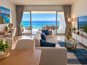 A modern living room with a large sofa, a dining area, and a balcony with a view of the ocean.