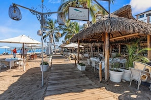 A beachside restaurant with outdoor seating, a wooden deck, and thatched-roof structures.