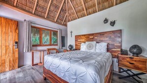 Een slaapkamer met een houten bed, een nachtkastje, een bureau, een stoel, een raam met gordijnen en een rieten dak.