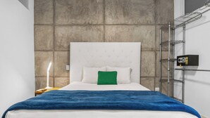 Habitación de hotel con cama, mesita de noche, lámpara, almohada verde y una cámara de seguridad en la pared.