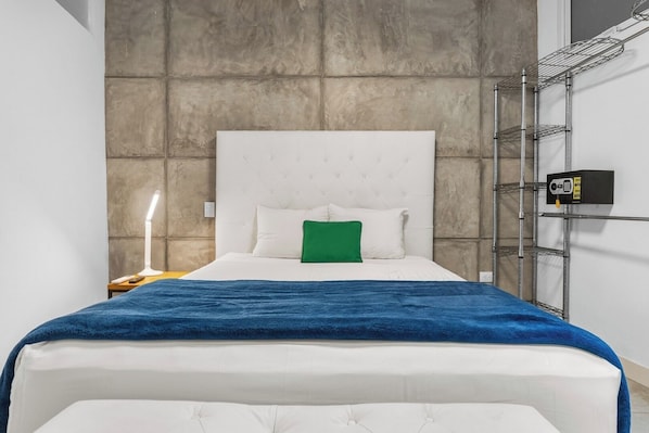 A hotel room with a bed, a nightstand, a lamp, a green pillow, and a security camera on the wall.