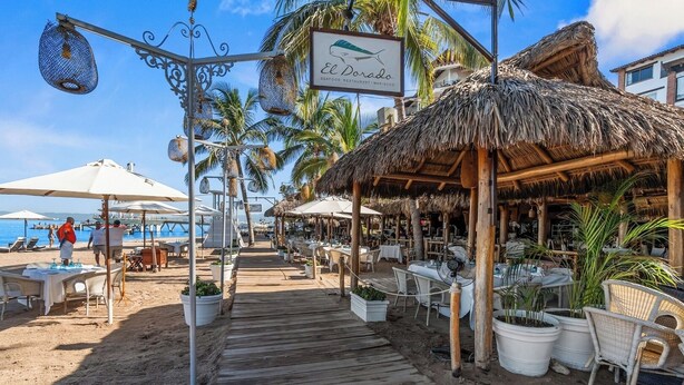 Un restaurante frente a la playa con terraza, una cubierta de madera y construcciones de techo de paja.
