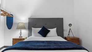 Een netjes opgemaakt bed met een grijs hoofdeinde, witte kussens en een blauwe plaid. Er zijn twee nachtkastjes, waarvan één met een lamp en de ander met een lamp op een statief.