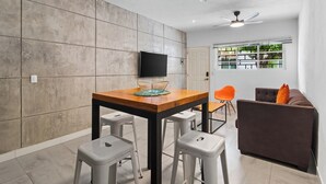 A modern kitchen with a wooden table, two bar stools, a TV mounted on the wall, and a window with a view of greenery. - Simple loft in a romantic area + Wi-Fi + beach (Puerto Vallarta)