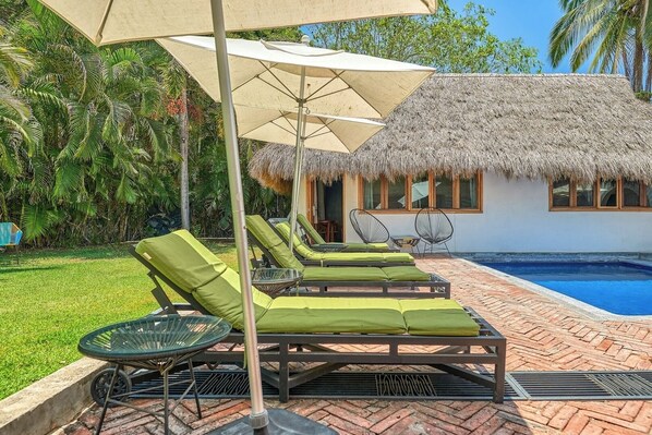 Un espace aménagé au bord de la piscine, avec des chaises longues, un bâtiment à toit de chaume et une piscine.