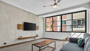A modern living room with a flat-screen TV mounted on a textured wall, a wooden coffee table, a gray sofa with a colorful pillow, and a large window with a view of a brick building. - Prime location in the Romantic Zone + beach (Puerto Vallarta)