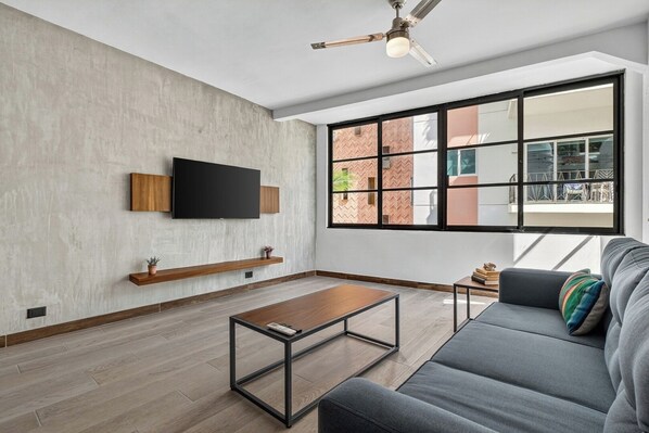 A modern living room with a flat-screen TV mounted on a textured wall, a wooden coffee table, a gray sofa with a colorful pillow, and a large window with a view of a brick building.