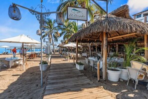 Un restaurante frente a la playa con mesas al aire libre, un paseo de madera y estructuras de techo de paja.