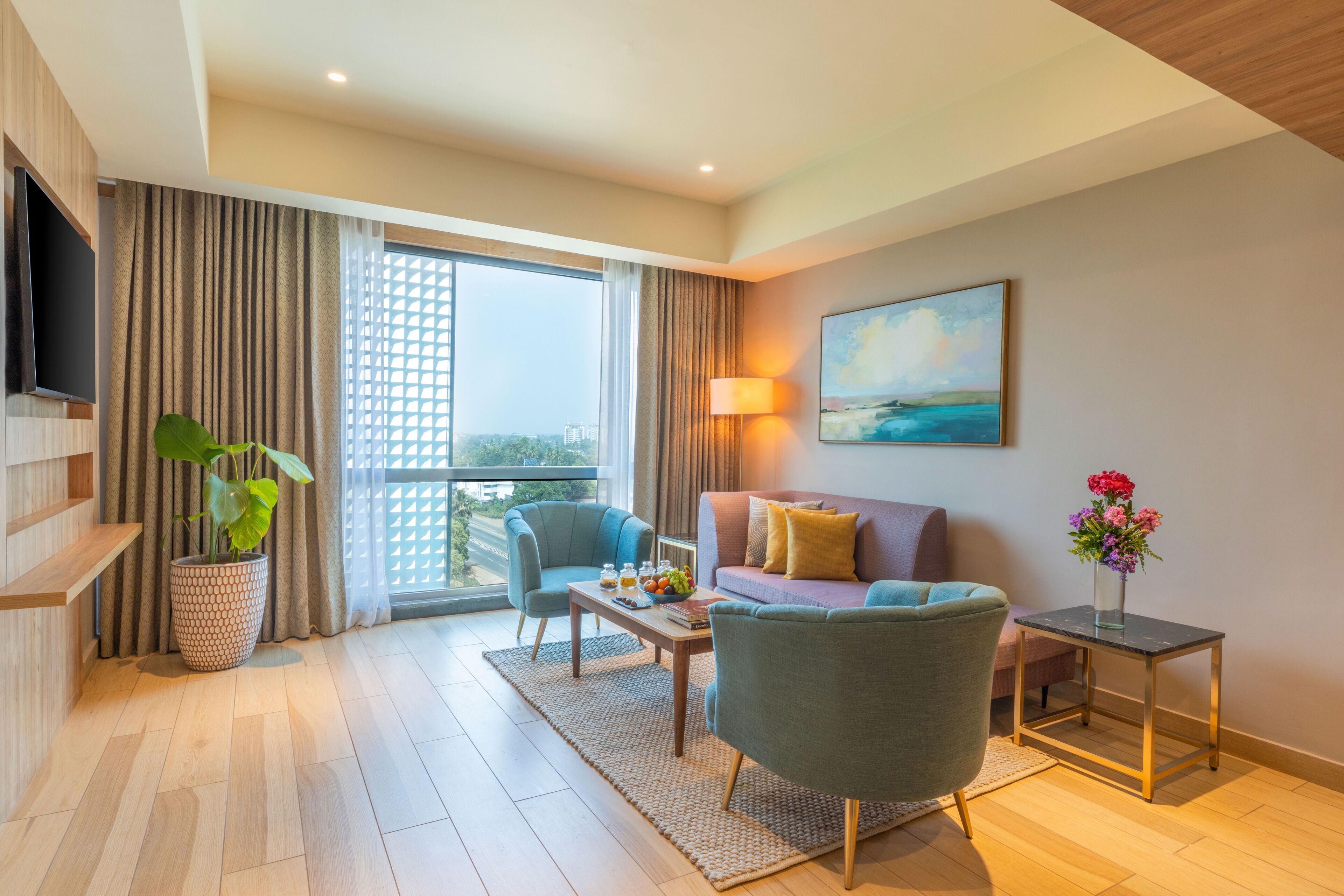 Quarto de hotel moderno com uma janela grande, televisão de tela plana, sofá, mesa de centro e planta em vaso.