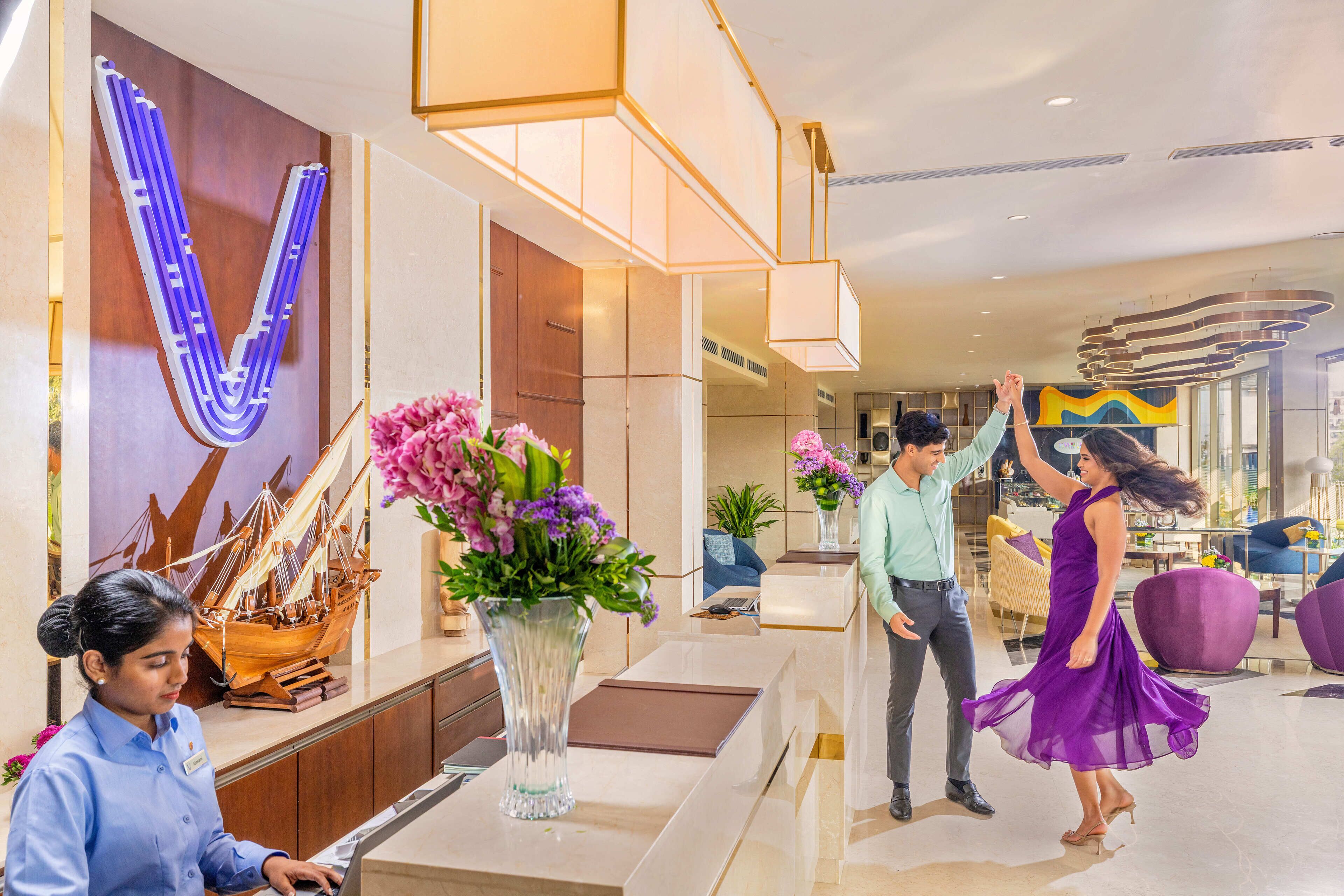 A hotel reception area with a large V-shaped light installation, a reception desk, a vase with flowers, and a ship model.