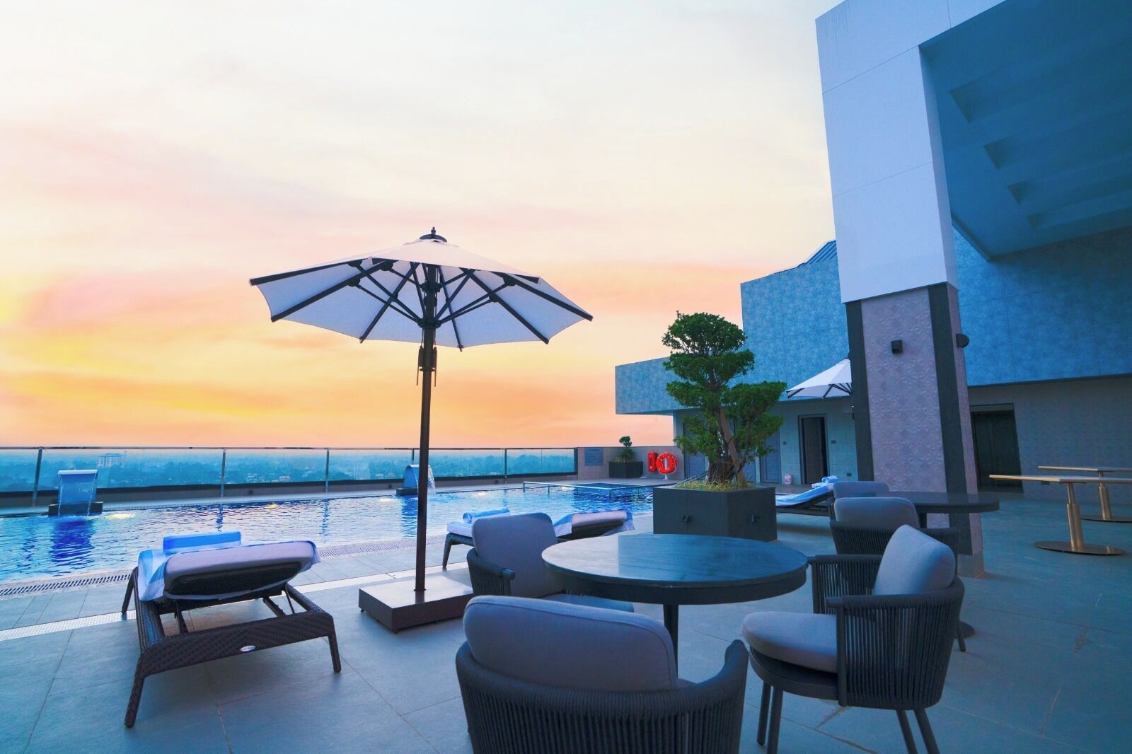 Un espace piscine aménagé sur le toit, avec des chaises longues, une table et un parasol, offrant une vue sur le paysage urbain au coucher du soleil.