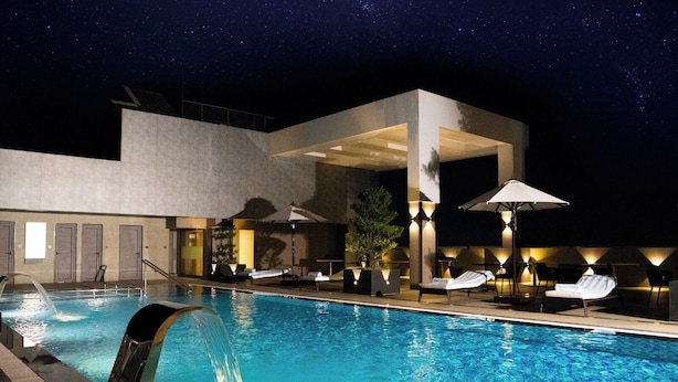 A swimming pool with lounge chairs and umbrellas, surrounded by a modern building with a covered area. - Vivanta Kochi Aluva (Aluva)